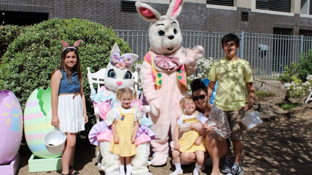 Four children and one adult are standing next to two people dressed up in Easter Bunny costumes posing with their Easter baskets.