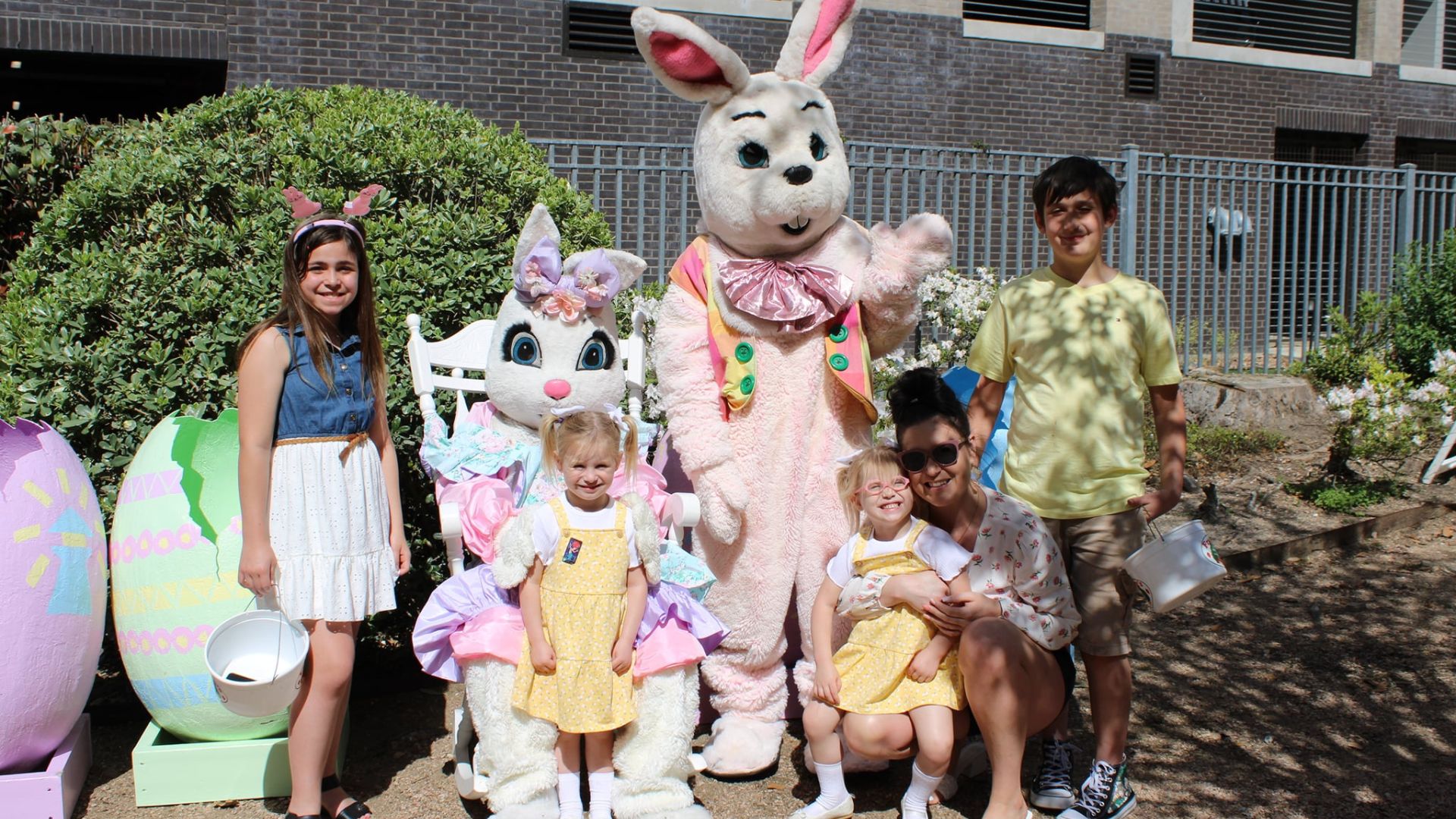 Four children and one adult are standing next to two people dressed up in Easter Bunny costumes posing with their Easter baskets.
