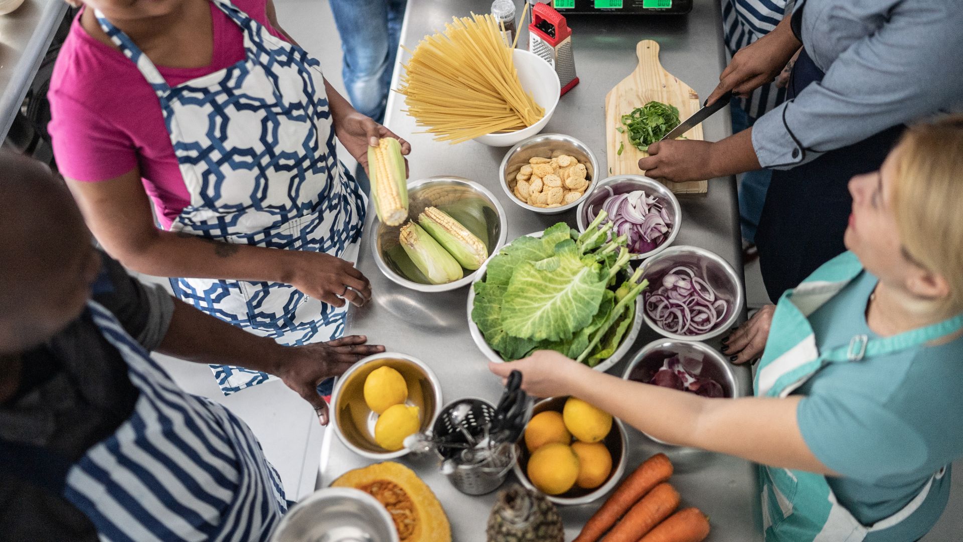 Four people standing around a counter with food prepped in multiple different bowls. There's pasta, corn, lemons, other fruits and vegetables, and one person is chopping up herbs.