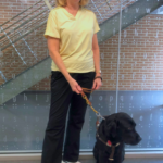 A volunteer standing with her guide dog in front of the braille wall at LOH.