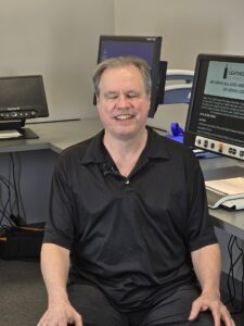 Photo of Steven seated in front of several computer monitors with a happy smile.