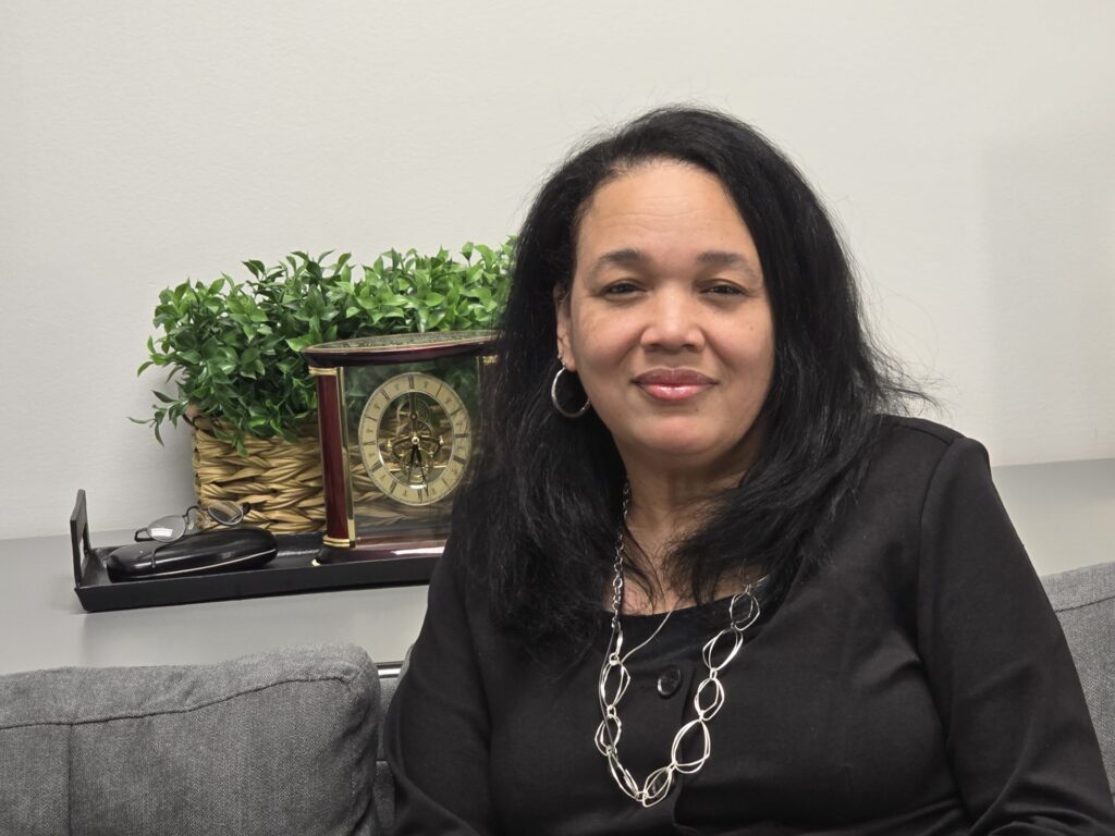 Photo of Victoria Hillard seated on a gray couch with a beautiful smile. A green plant and clock appear on a shelf behind her.