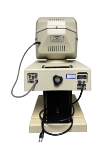 A cream-colored Optelec with a top compartment featuring ventilation slots and a warning label, connected by a gray wire to a lower control panel with various ports, a dial, and a black power cord plugged into a base.