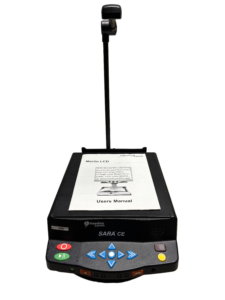A SARA CE device with a users manual open on top, a camera on a stand above it, and control buttons on the front.